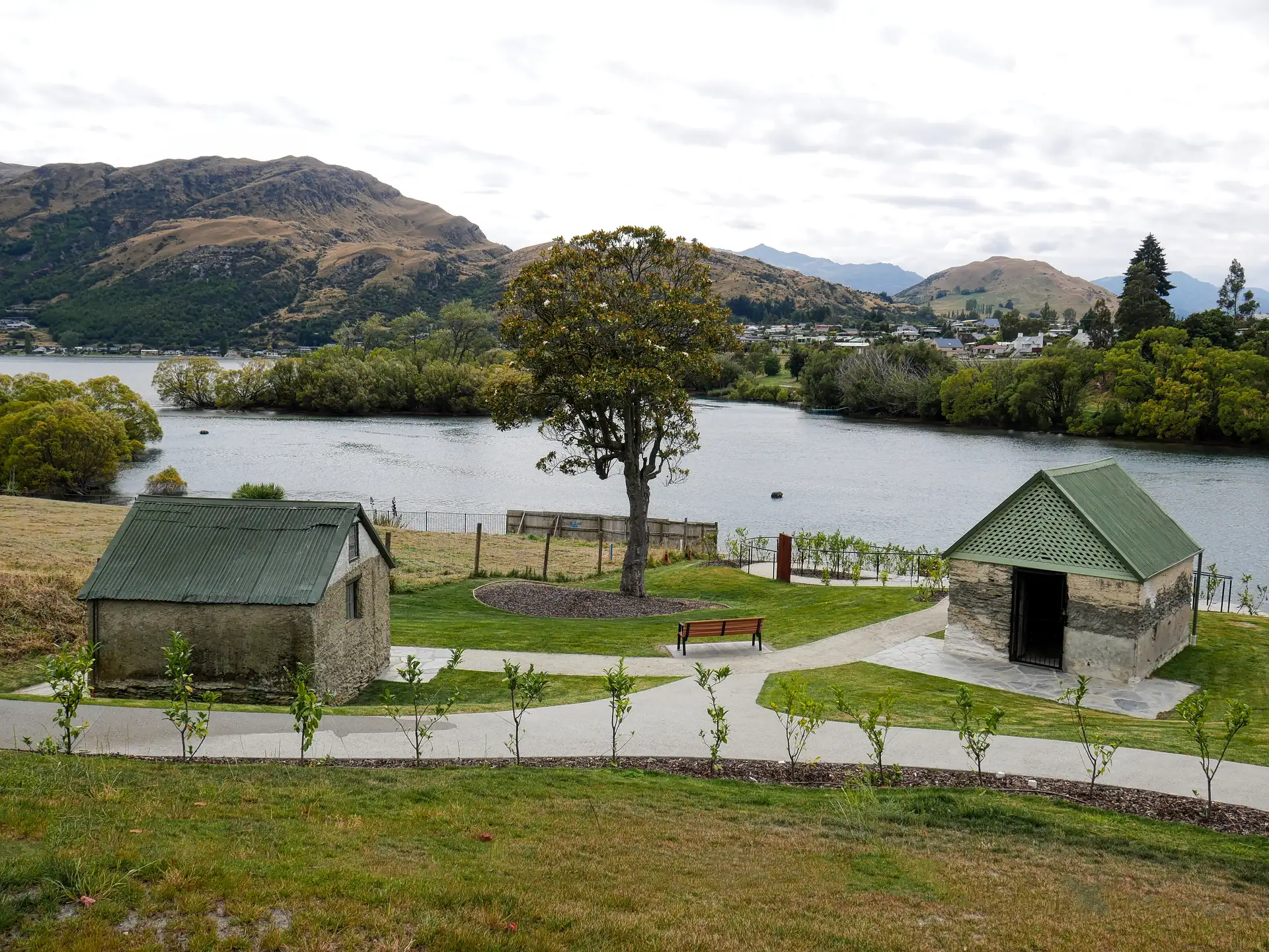 Two small stone outbuildings with green corrugated roofs at Rees Homestead Park, with paths, a bench and a single large tree above the water