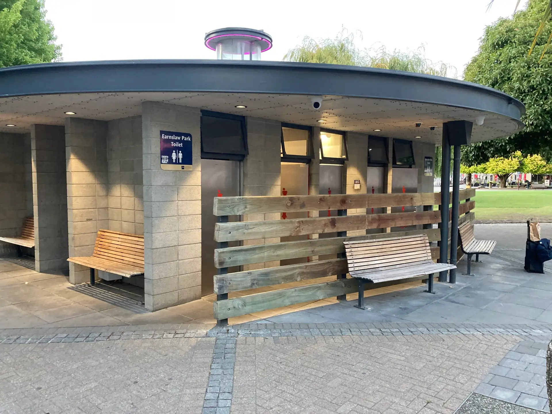 The circular Earnslaw Park toilet block on the Beach Street edge of the park, with its signposted entrance, row of cubicle doors and benches under a wide oval canopy