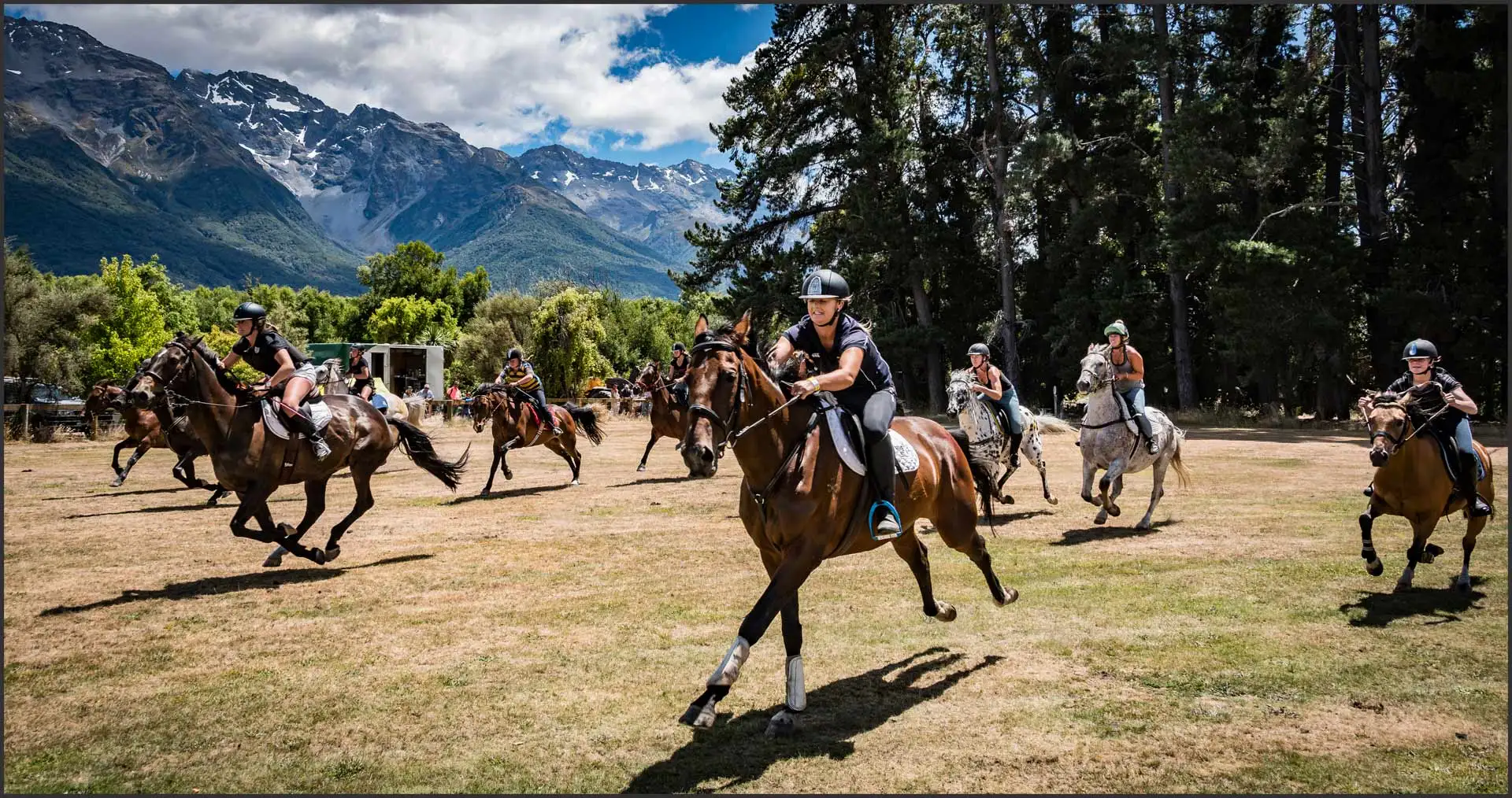 Riders galloping across the grass track at the Glenorchy Races, with beech trees and the snow-streaked Humboldt Mountains behind