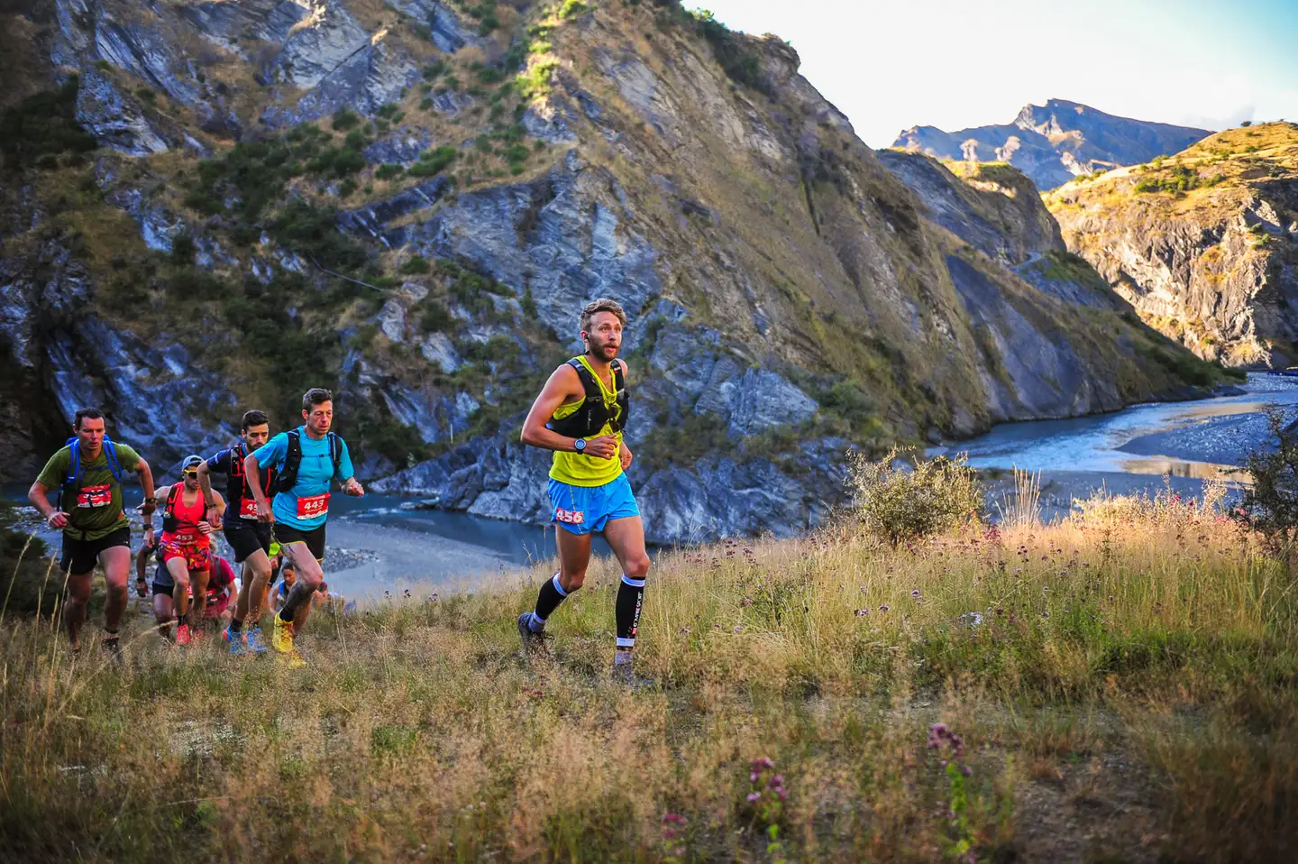Trail runners climbing a tussock track above the Shotover River on the Shotover Moonlight course, with schist bluffs behind them