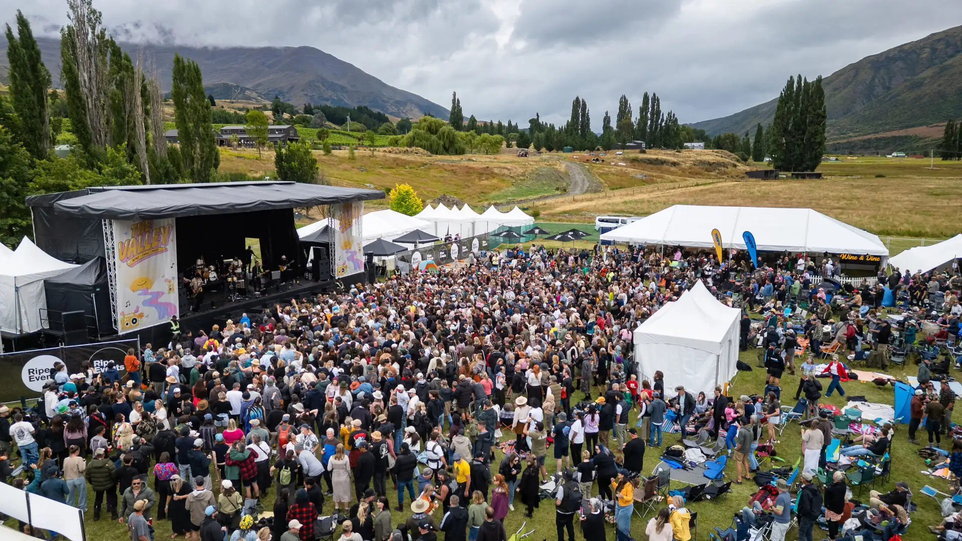 Aerial view of the crowd packed in front of The Valley festival stage on a grassy Gibbston paddock, with wine marquees, deck chairs and the Kawarau Gorge hills behind