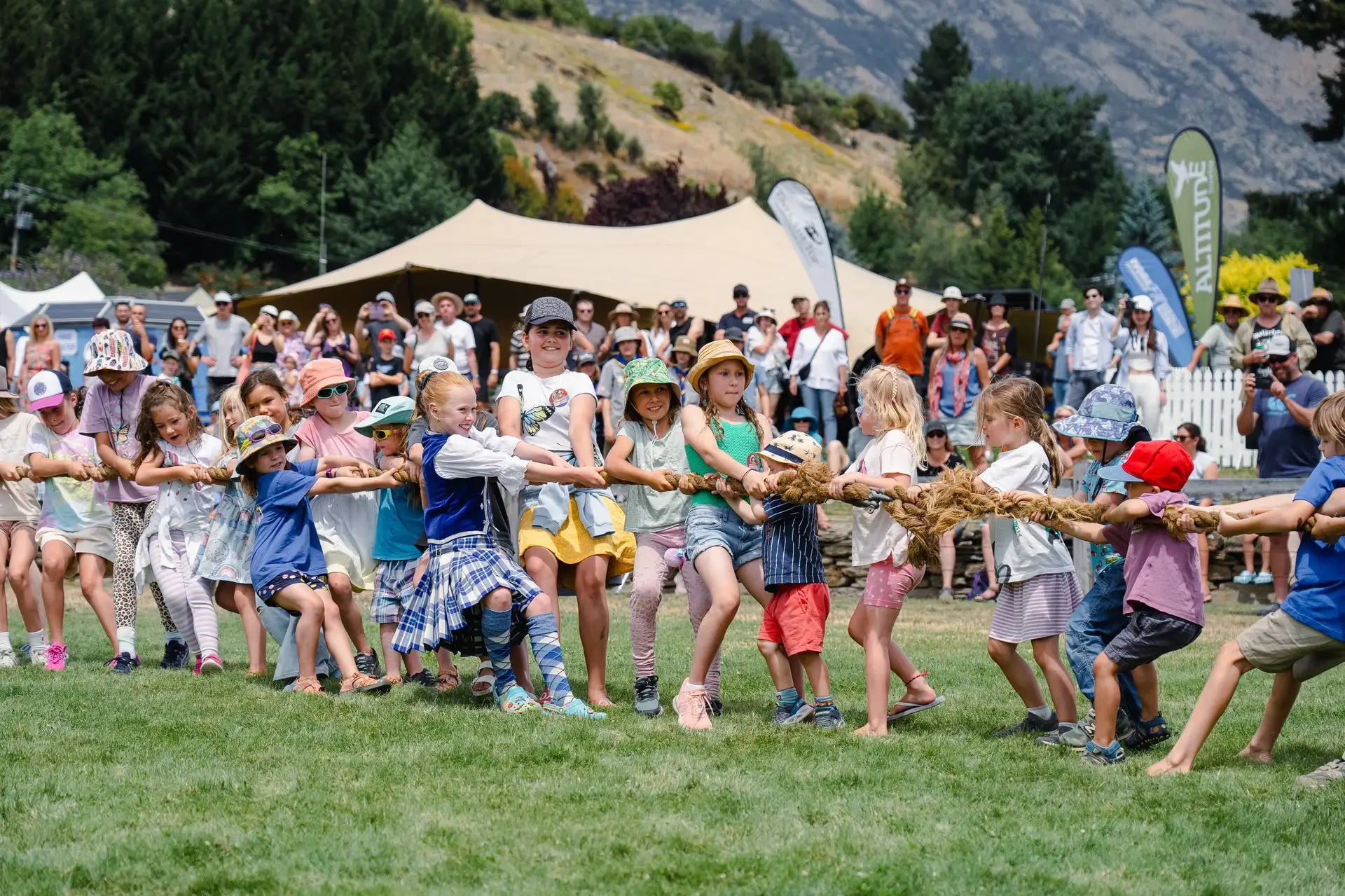 Children in sunhats hauling on a rope during the tug of war at the Lake Hayes A&P Show, with the showgrounds crowd and hills behind