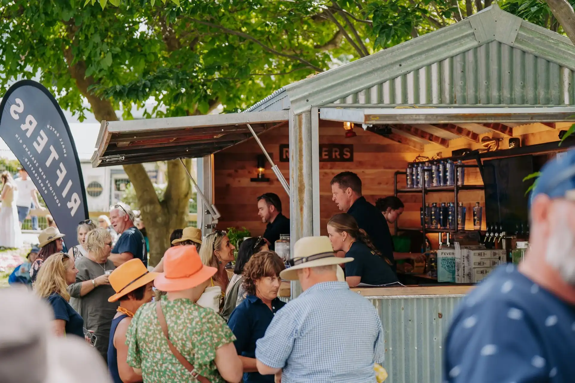 Festival-goers in sun hats queuing at a corrugated-iron gin bar shack under leafy trees at Gin Fest Arrowtown