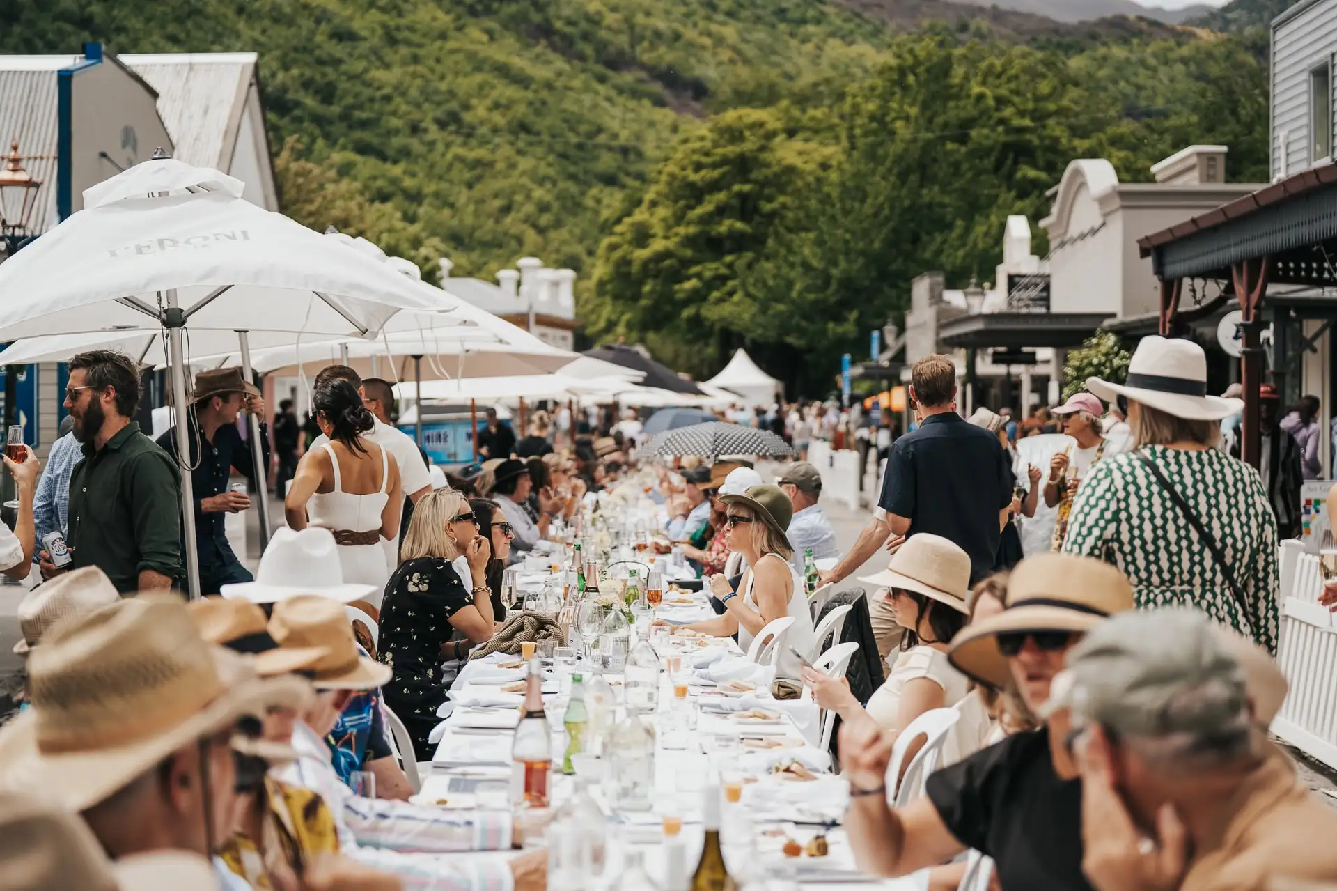 Diners in sun hats seated along one long white-clothed table running down Arrowtown's main street under umbrellas, with the bush-covered hills behind