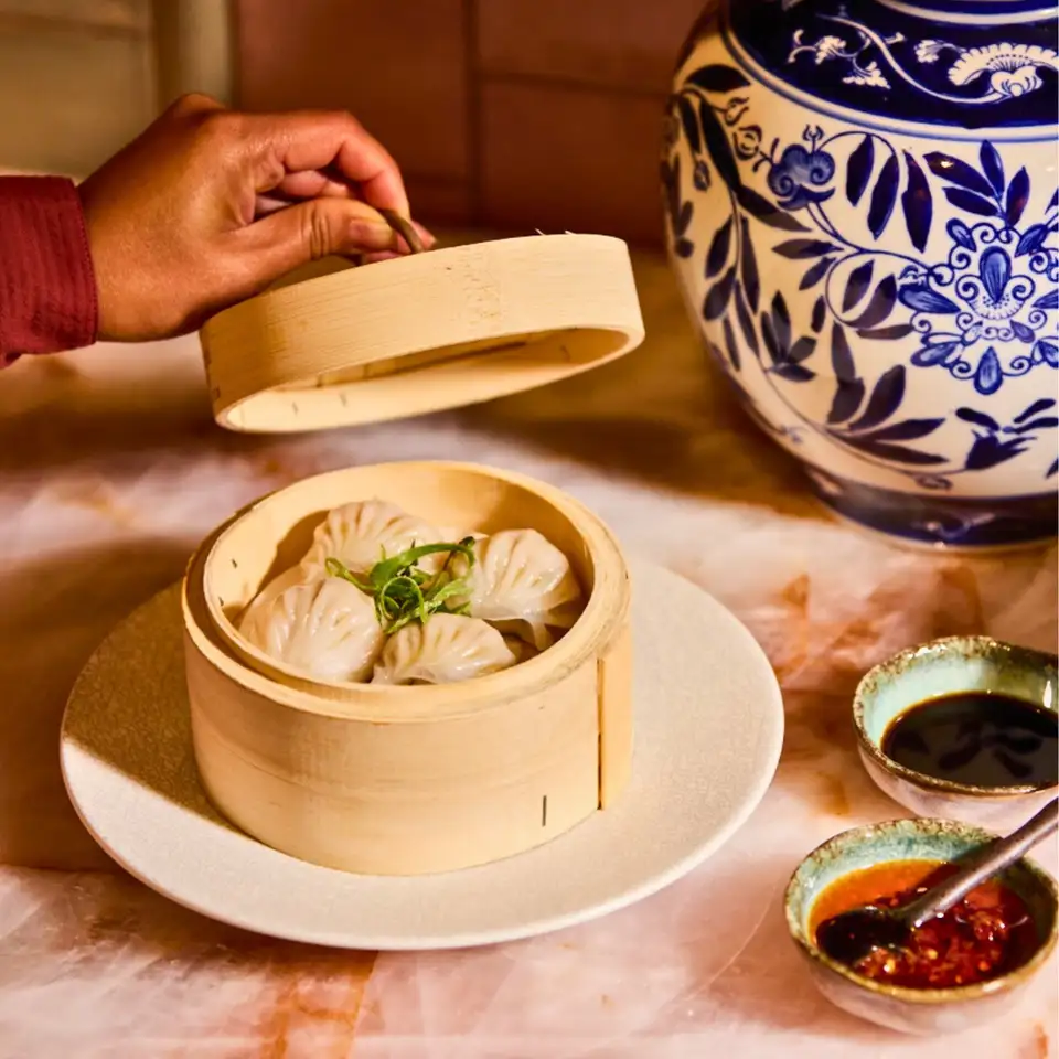 A bamboo steamer of prawn dumplings on a plate at Billy's, with soy and chilli dipping sauces and a blue and white ginger jar alongside