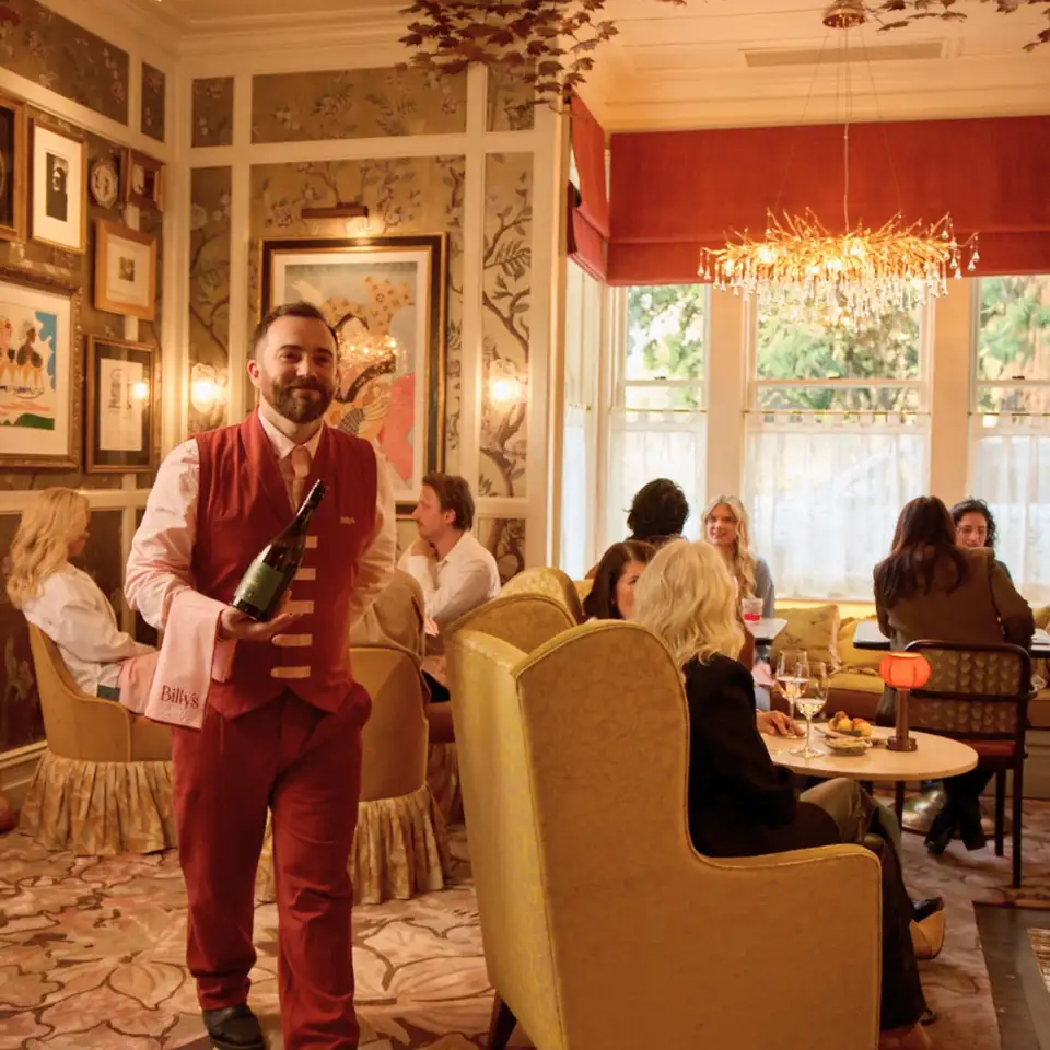 A waiter in a rust-red suit carries a bottle of wine through a floral-papered room at Billy's where guests sit in armchairs beneath a chandelier