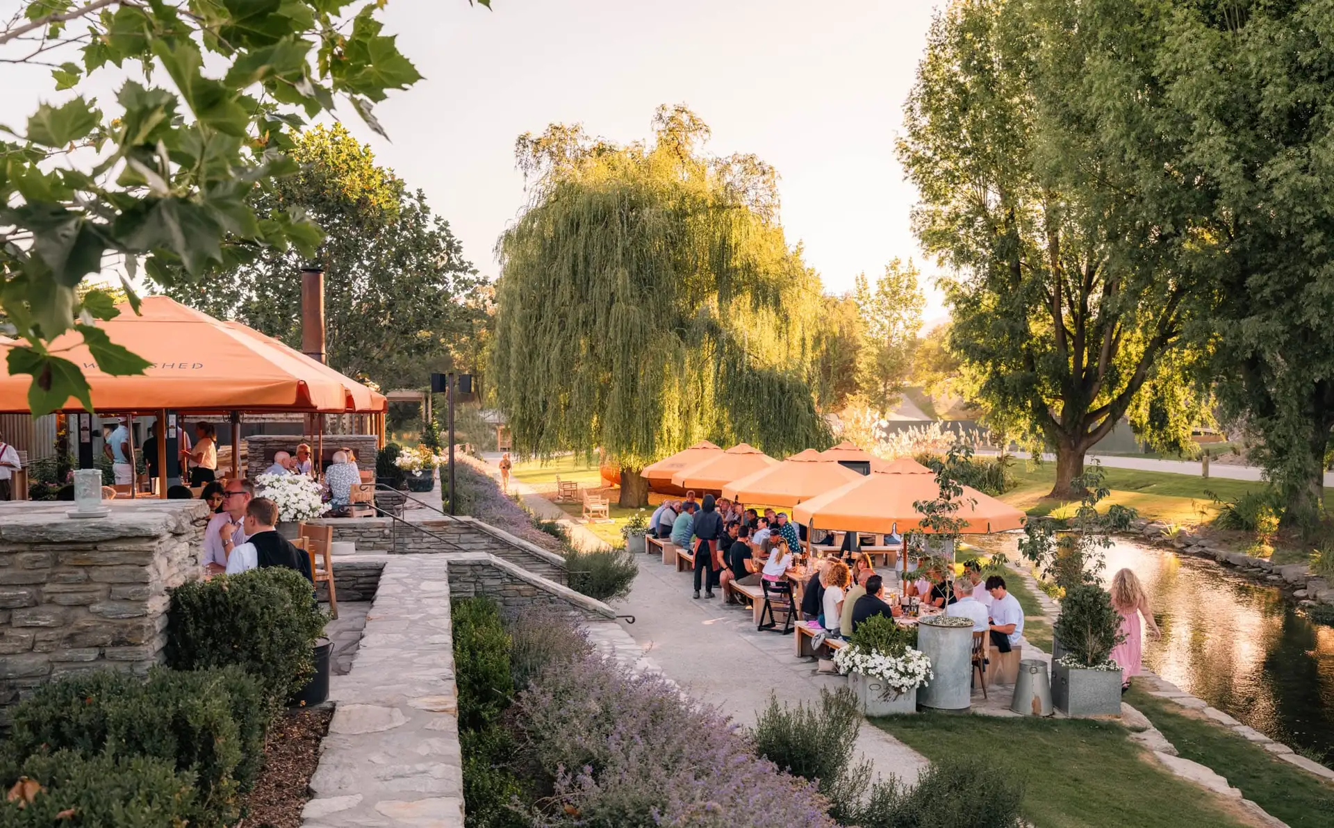 Evening at The Woolshed, with diners seated under orange umbrellas along the stone terrace and the lawn beside the stream at Ayrburn