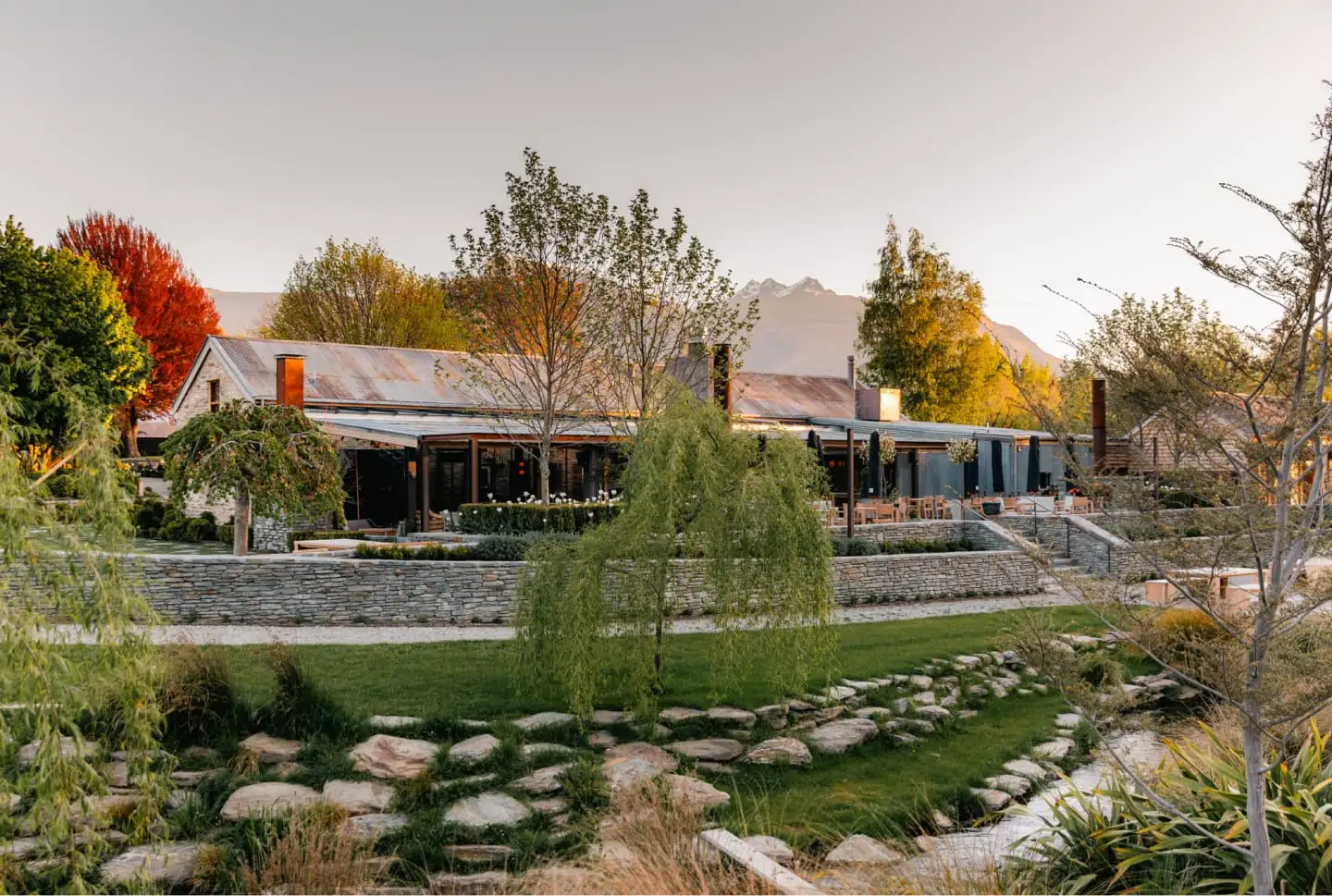 The stone and timber Woolshed building at dusk, with terraced lawns and stone walls in front and the mountains behind