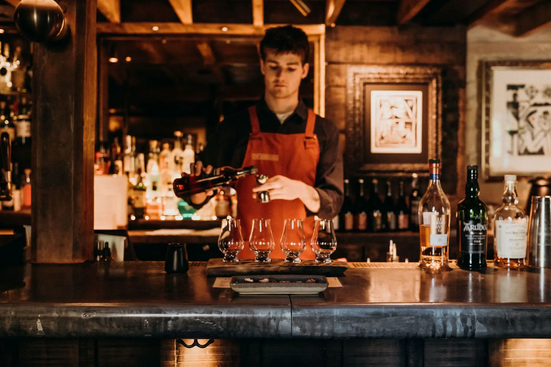 A bartender in an orange Woolshed apron pours a flight of whisky behind the timber bar