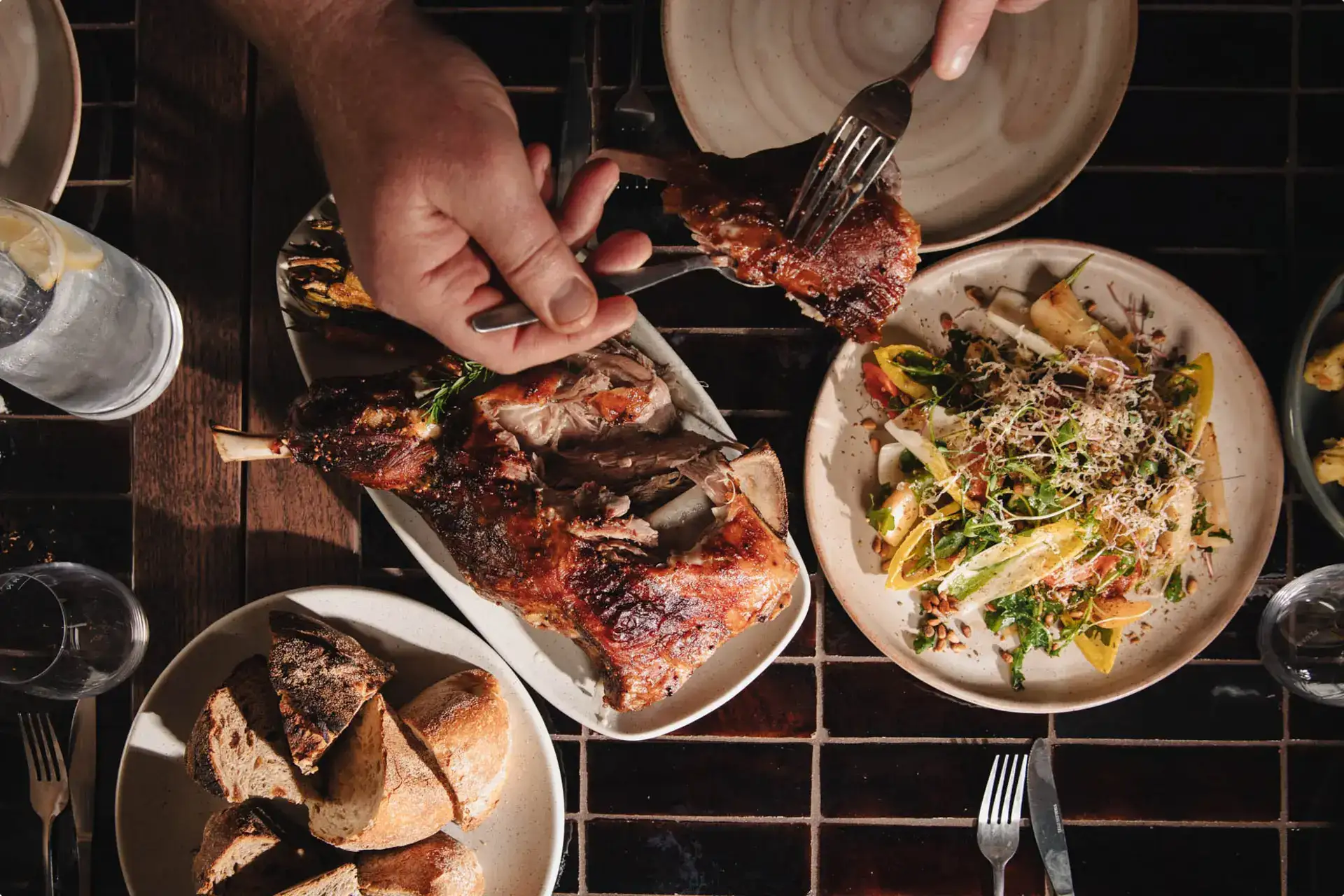 Overhead view of shared plates on a tiled table, with roast meat being carved onto a plate beside a salad and a bowl of sliced bread