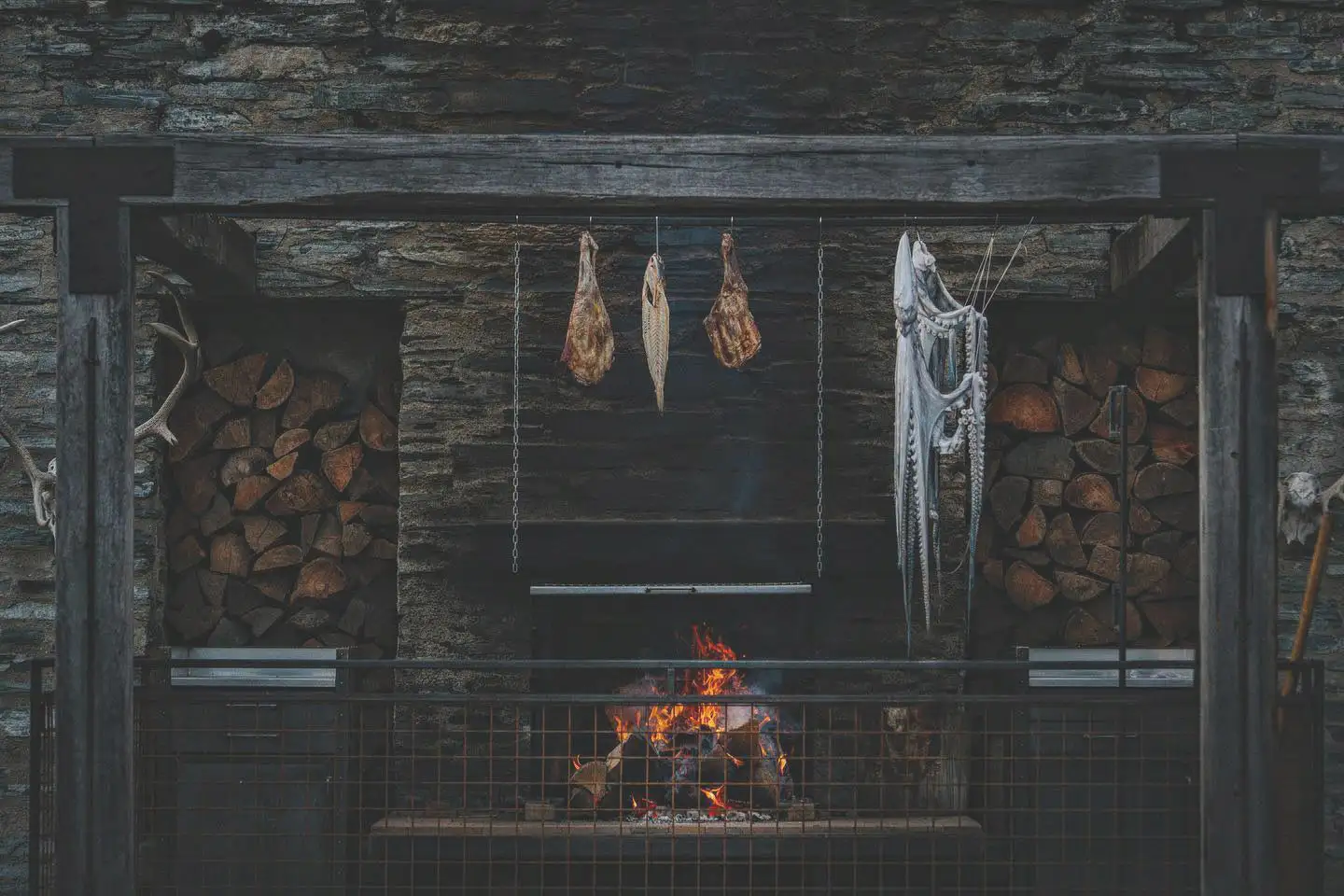 The open fire in the Amisfield kitchen, with cuts of meat and fish hung to cure above the flames between two stacked woodpiles