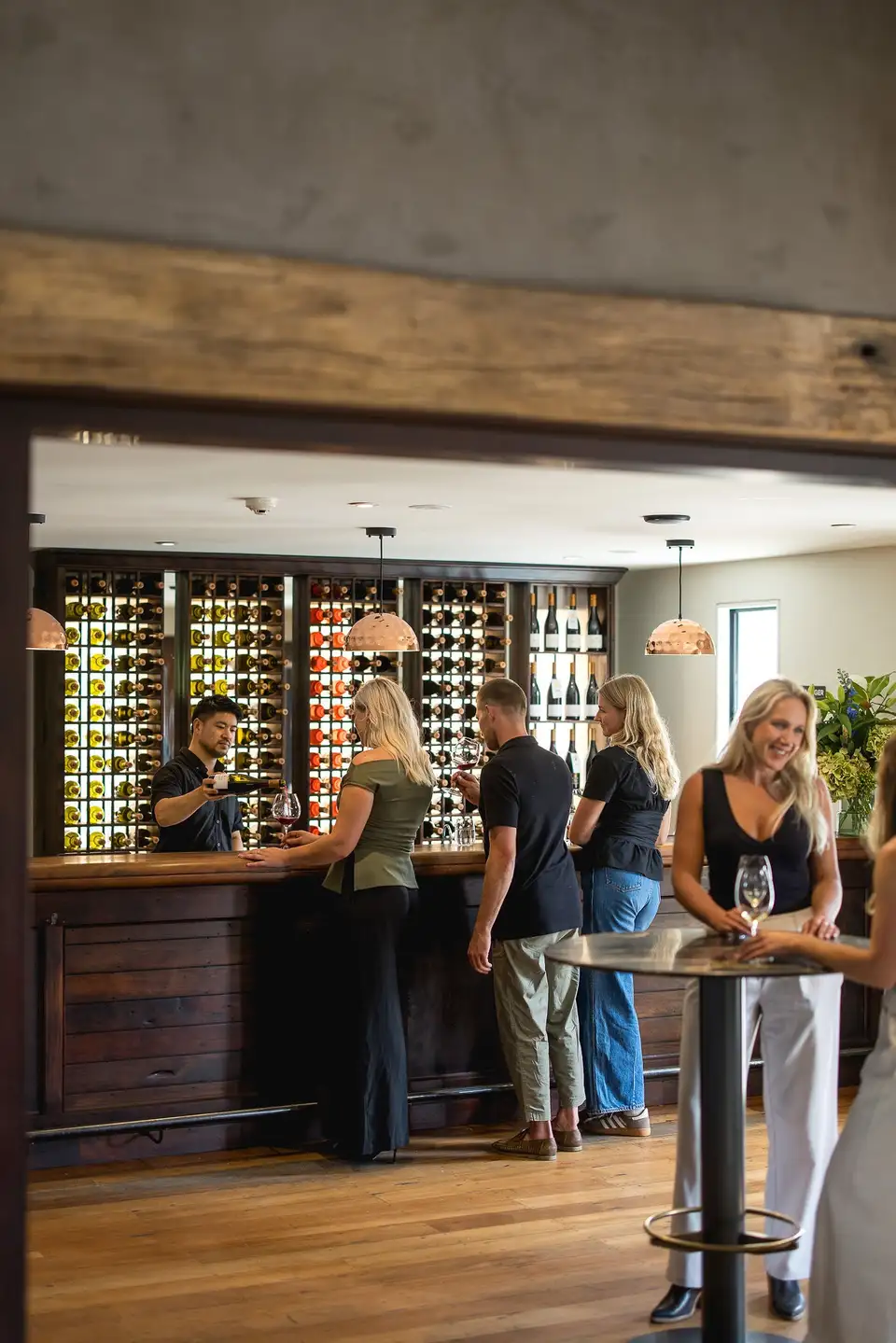Guests tasting wine at the timber bar of the Amisfield cellar door, in front of a backlit wall of bottles