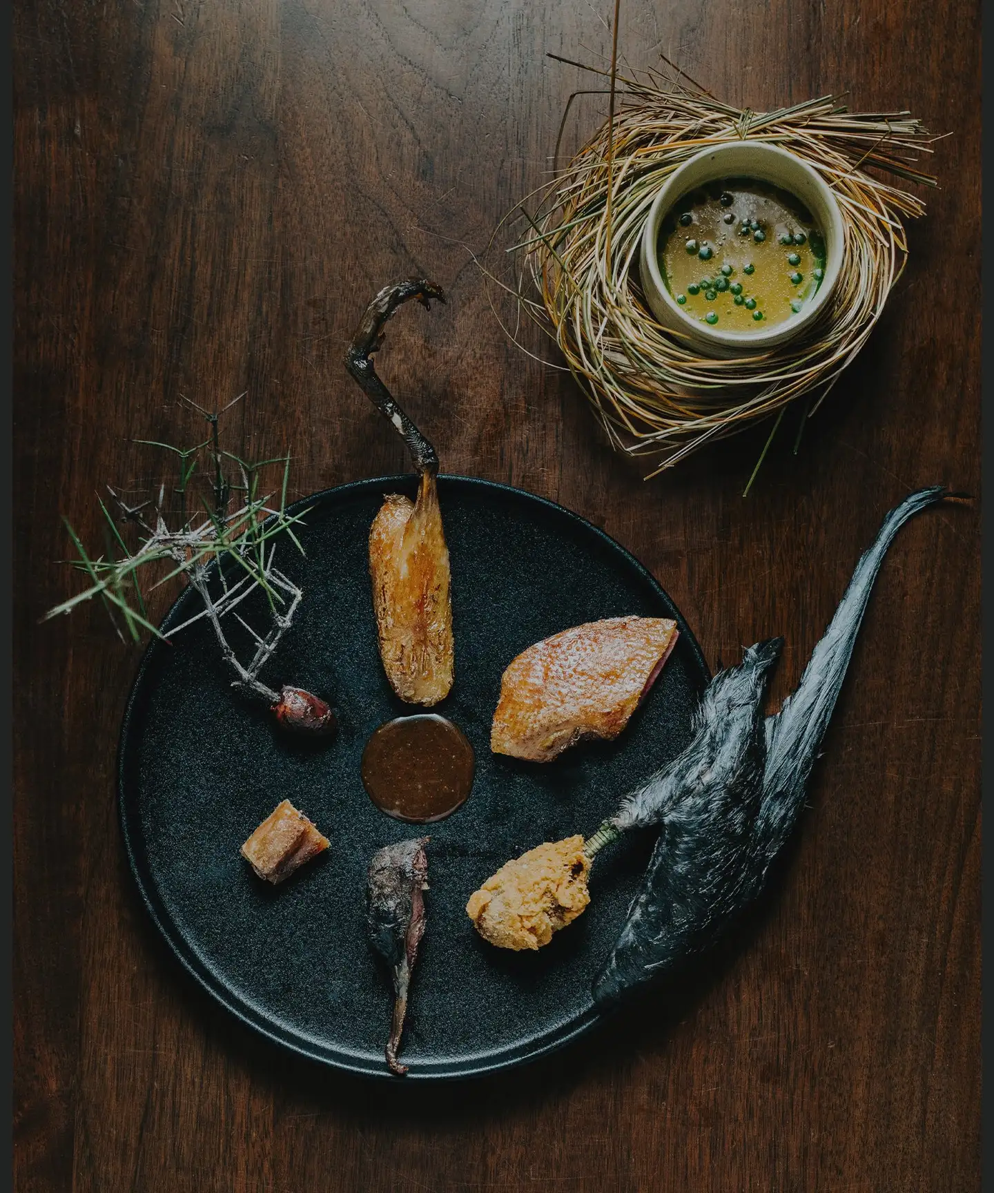 A game course at Amisfield plated on dark ceramic with roasted vegetables and a sauce, served alongside a broth set in a nest of dried grass