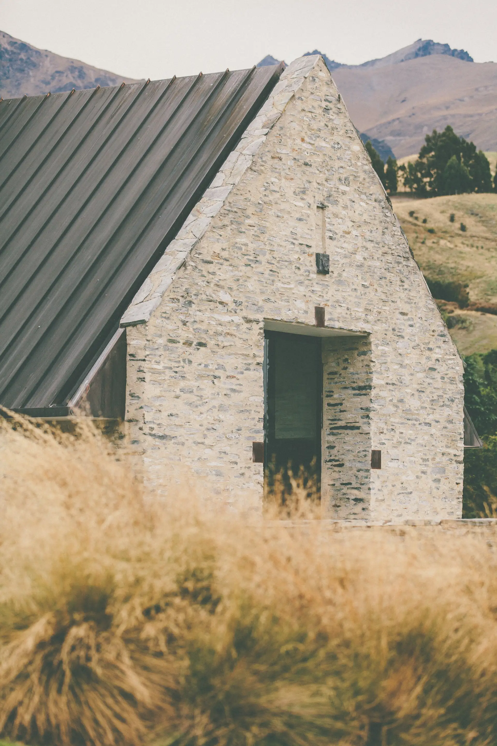 The stone gable end and dark pitched roof of the Amisfield building, with golden tussock in the foreground and the hills behind