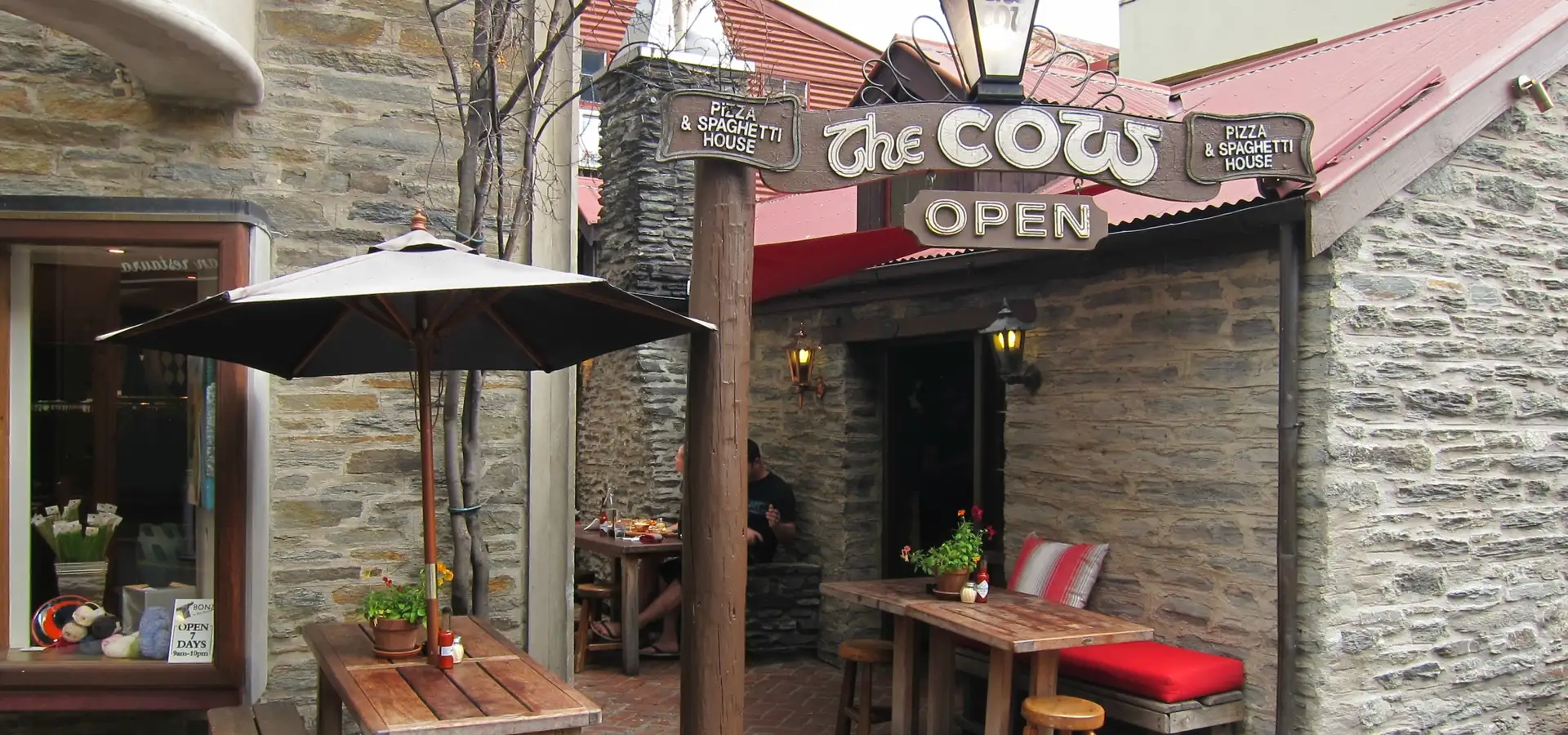 The stone frontage of The Cow on Cow Lane, with its hanging sign, lanterns and outdoor tables under an umbrella.