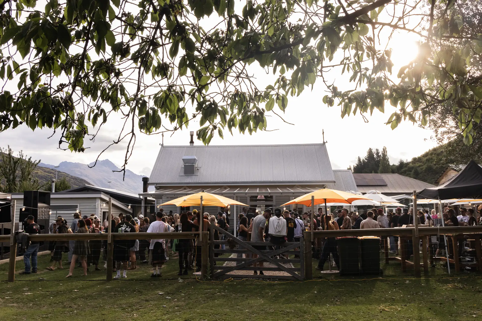 Diners gathered on the lawn at Sundays under yellow umbrellas on a summer evening, with the white weatherboard church building and mountains behind.