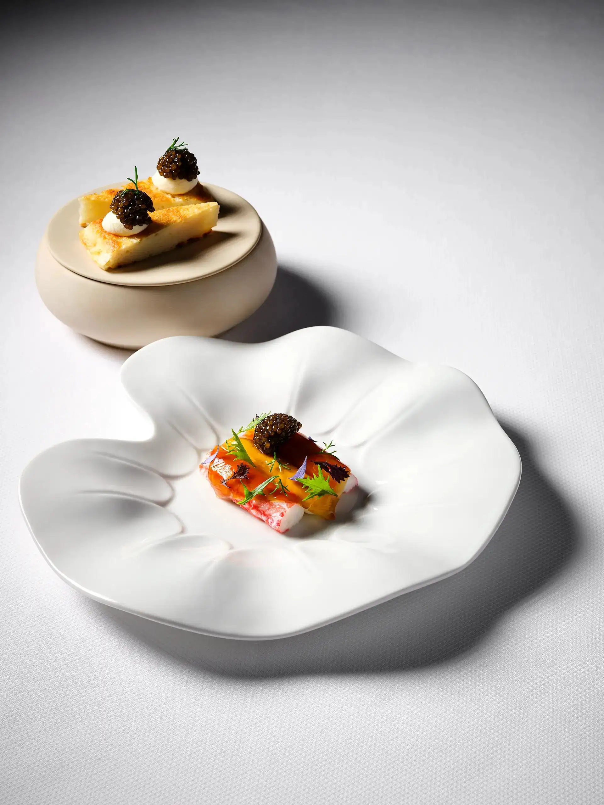 A king crab course topped with caviar on a petal-shaped white plate, with a lidded ceramic bowl behind.