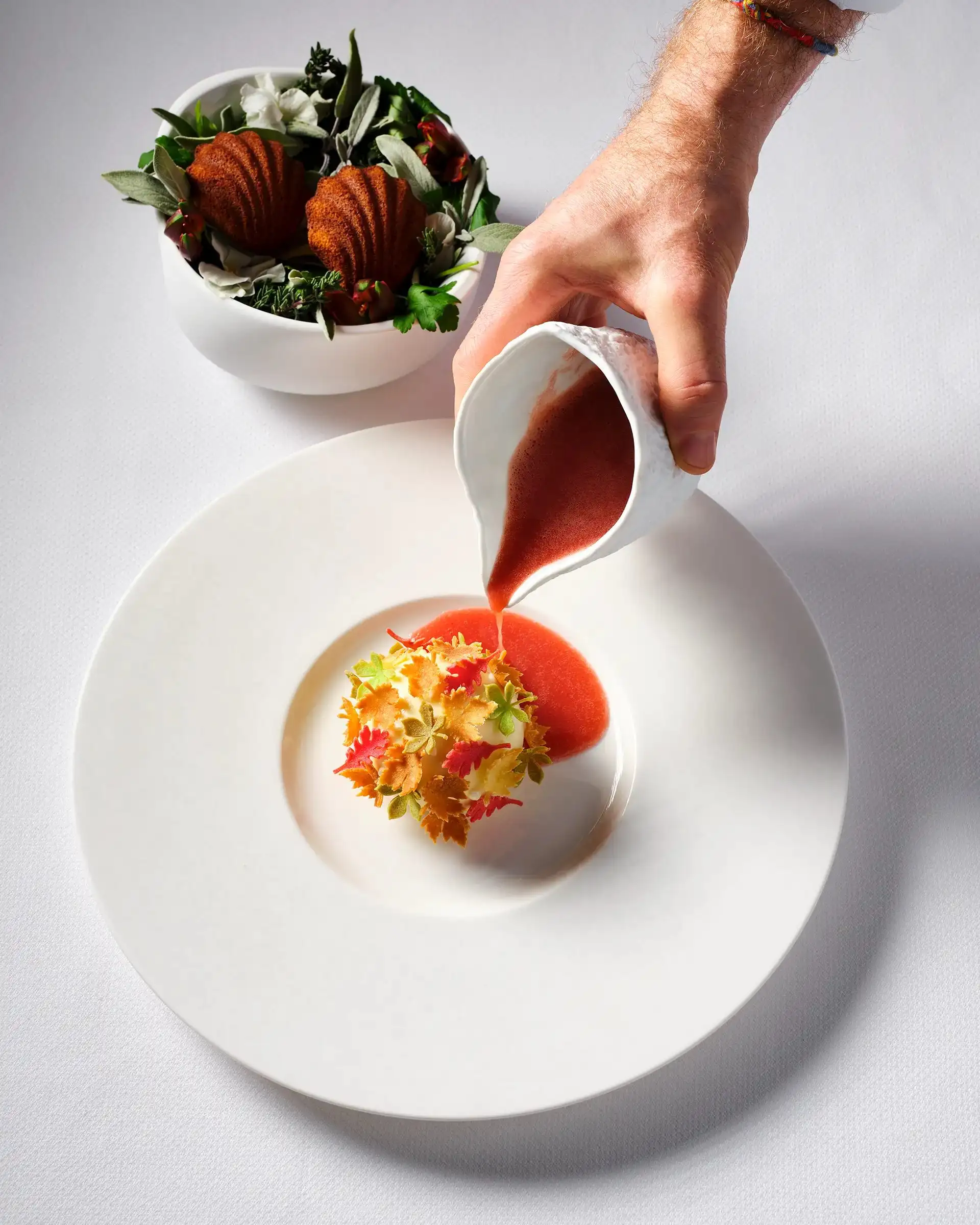 A hand pouring a red sauce from a small jug over a plated course at Essence, with a bowl of dressed leaves behind.