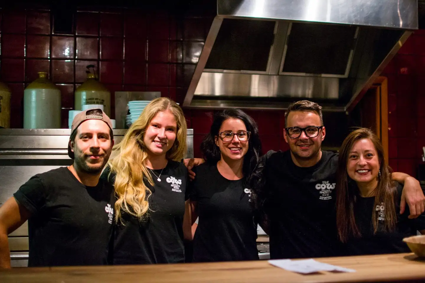 Staff behind the bar at The Cow, standing in front of the kitchen pass and the restaurant's red painted walls.