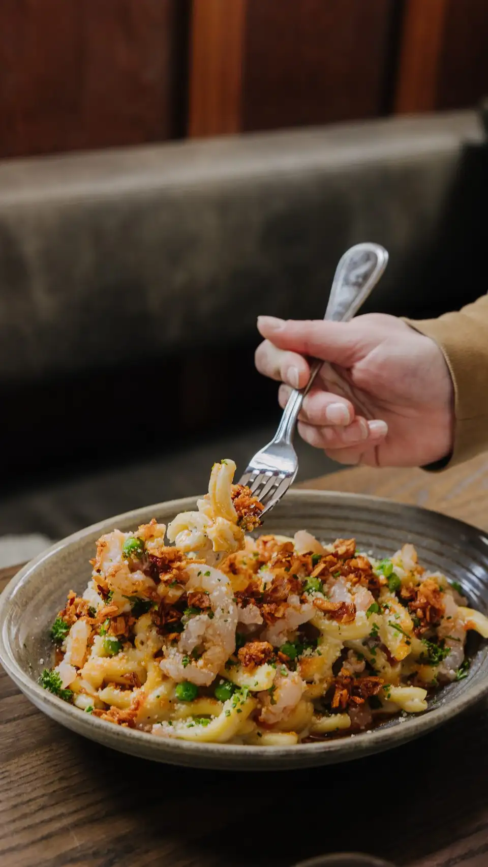 A hand lifting a forkful from a bowl of pasta with prawns, herbs and golden crumb on a wooden table at Sundays.