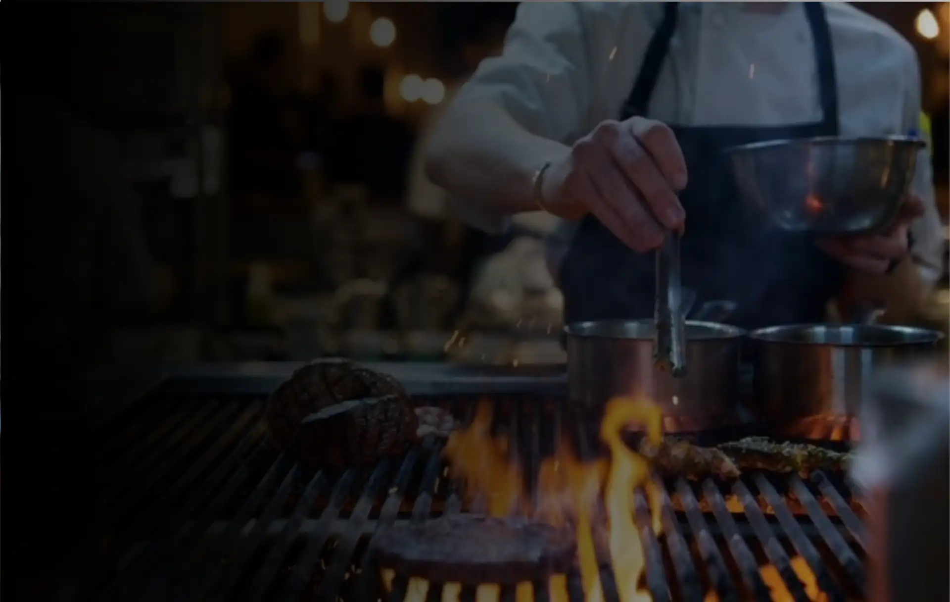 A cook working over the open wood-fired grill at Prospects, with steaks and flames below.