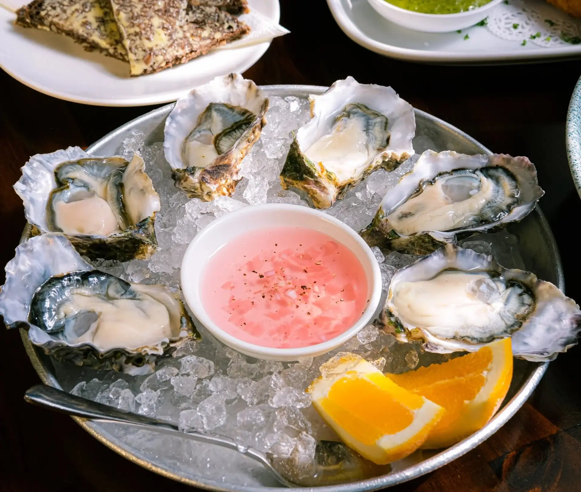 A platter of fresh oysters on ice with a bowl of pink mignonette and lemon wedges at Prospects.