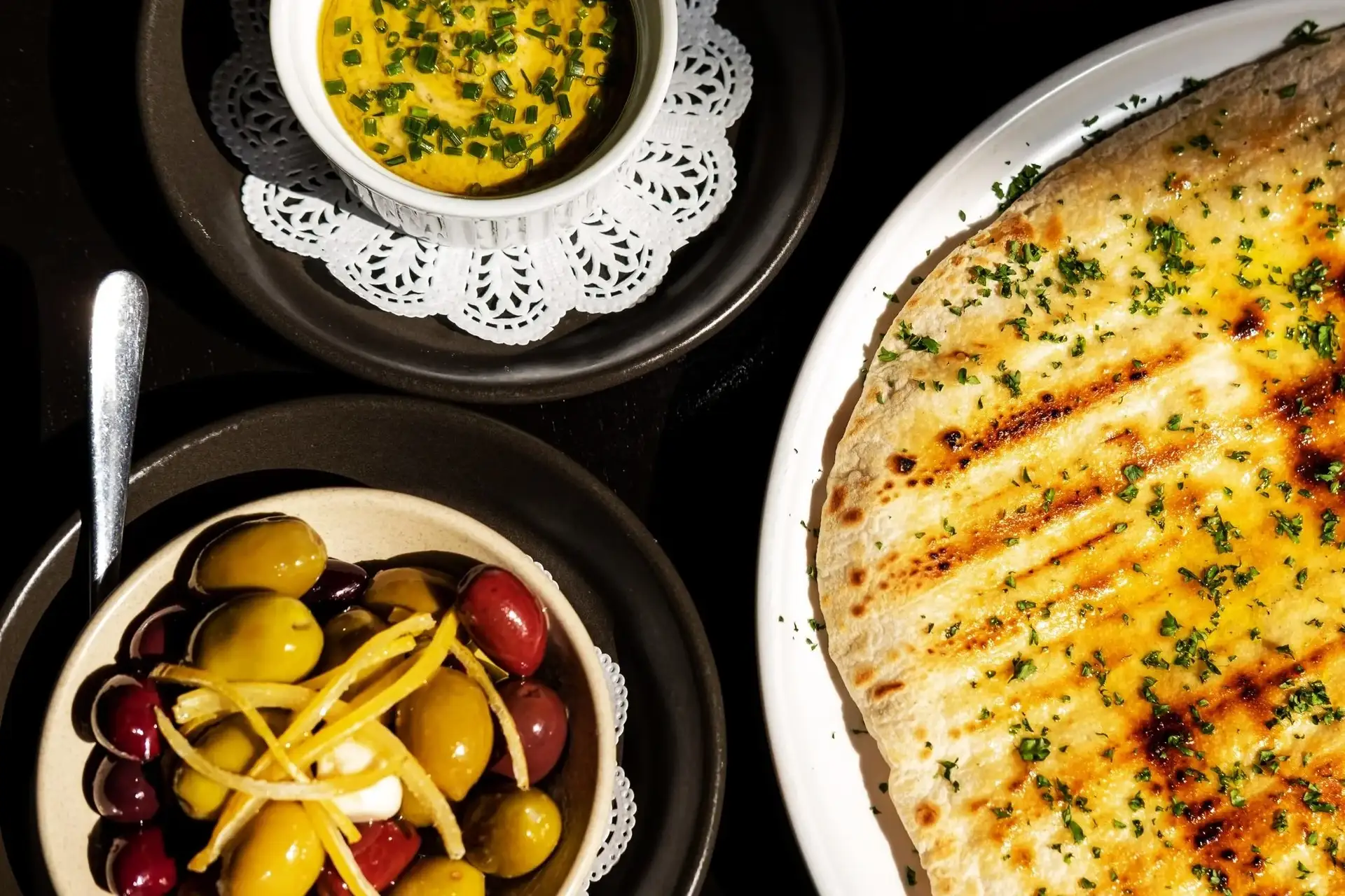 Grilled flatbread scattered with herbs, with bowls of olives and whipped butter, on a dark table at Prospects.