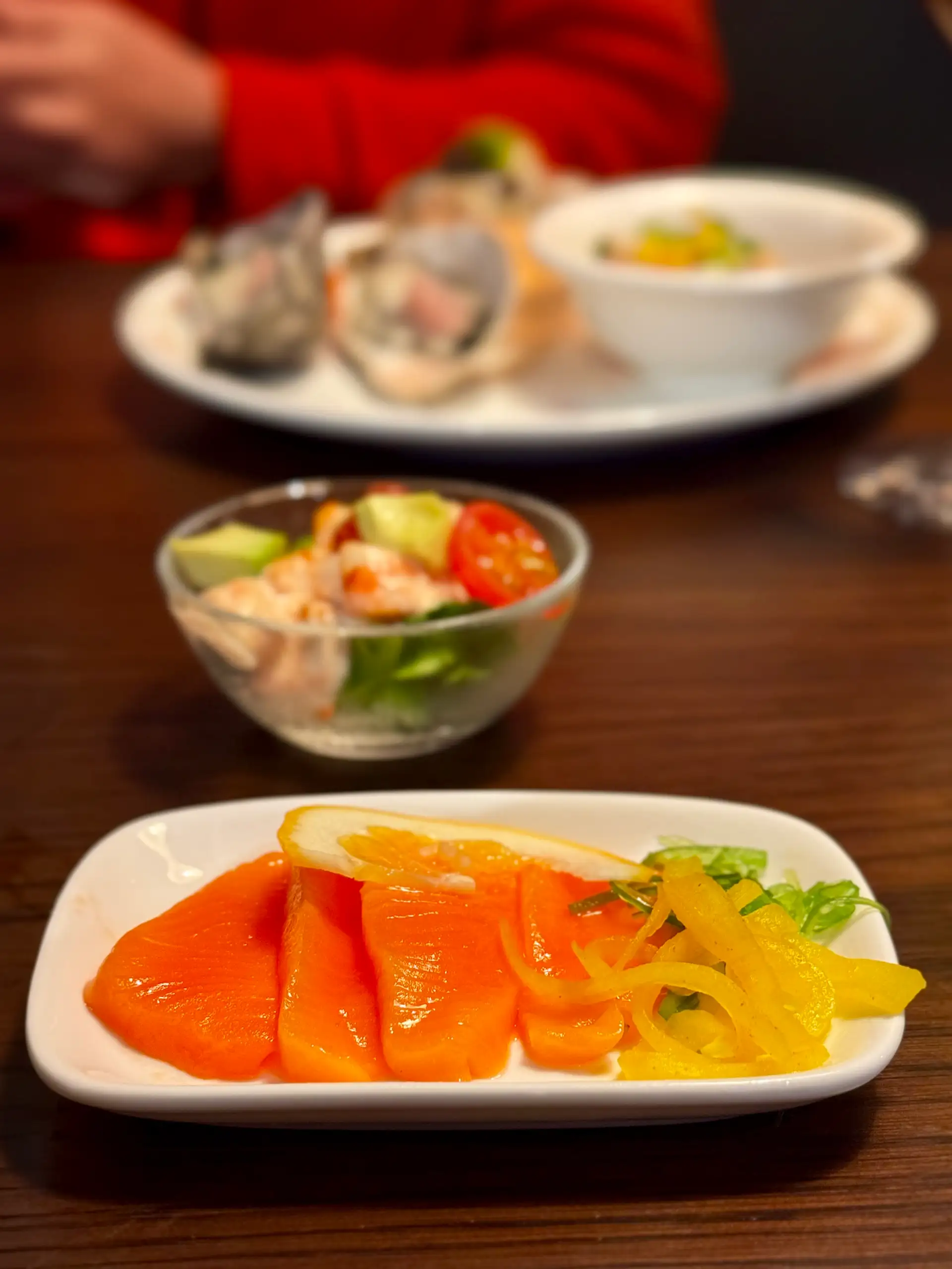 A plate of salmon sashimi with pickled peppers and lemon, with a prawn cocktail and a plate of oysters behind, at Prospects.
