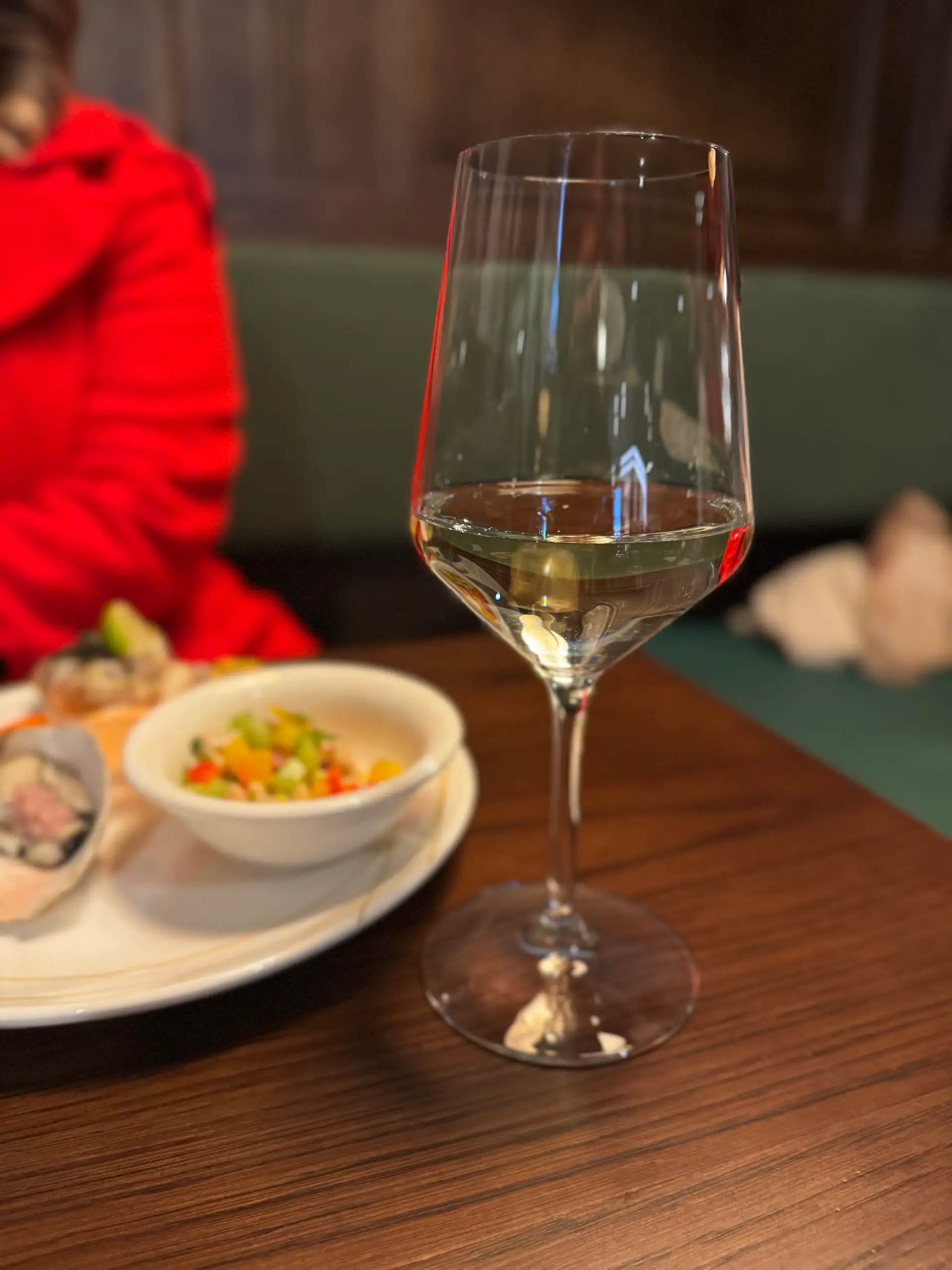 A glass of white wine beside a plate of oysters and a small bowl of ika mata on a timber table at Prospects, with a green banquette behind.
