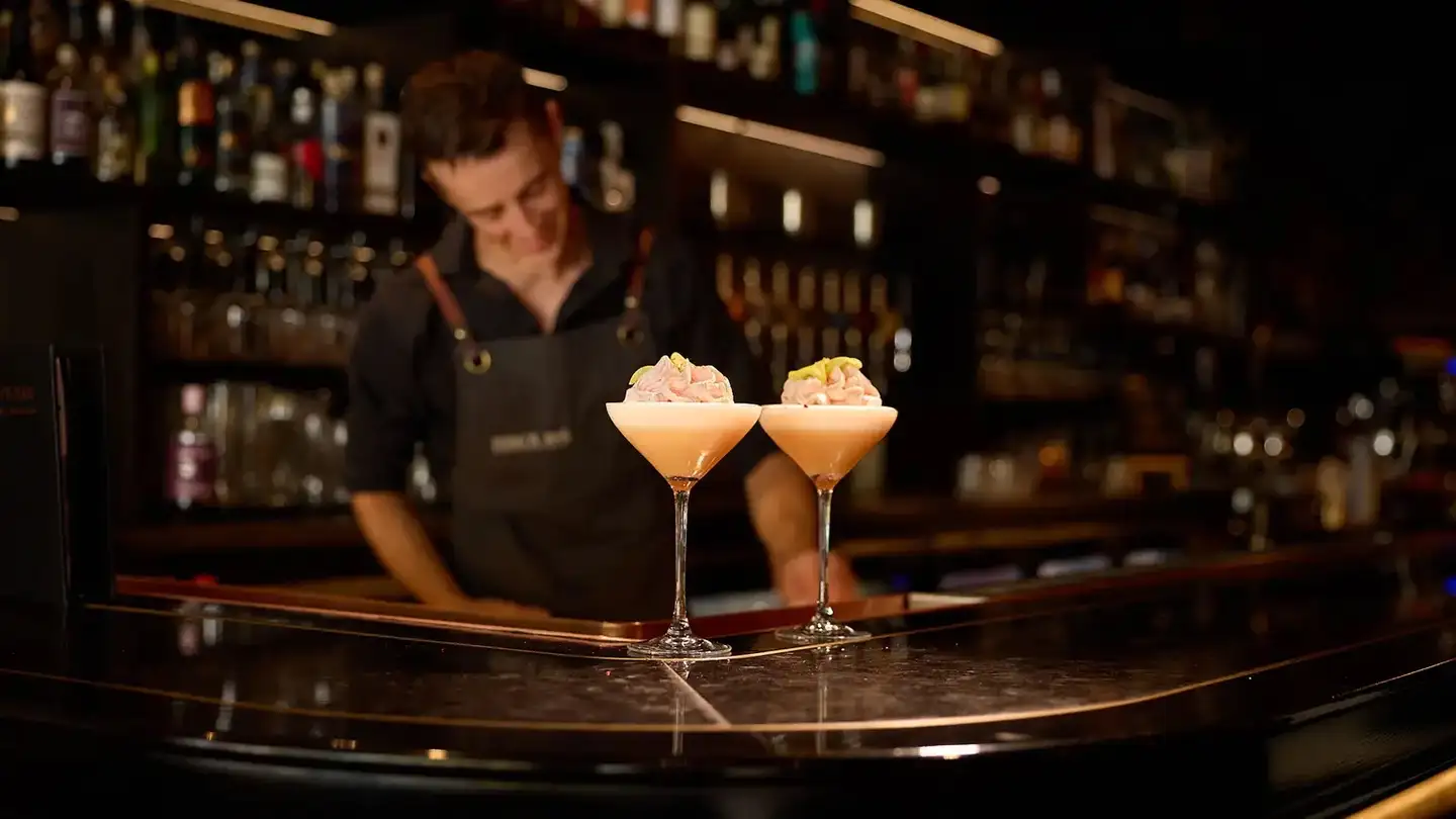 Two cocktails on the bar at Ferg's Bar, with a bartender and backlit spirits shelves behind